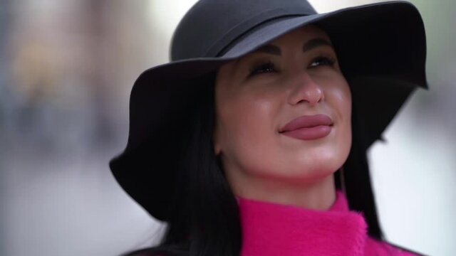 Close-up portrait of a pretty woman in a pink coat and black hat walking on a blurry background of a city street. Smiles.