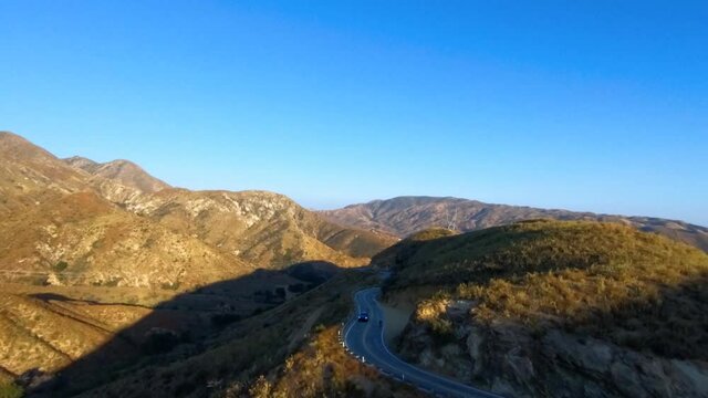 Aerial FPV, drone shot towards a blue suv car driving on a winding mountain road, sunny evening, in Los Angeles, California, USA - First person view