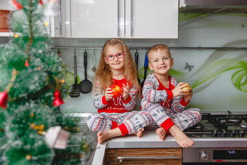 Obraz na płótnie Canvas Little laughing twins sit on the table in pajamas in the Christmas decorated kitchen with an Apple. Girl with glasses