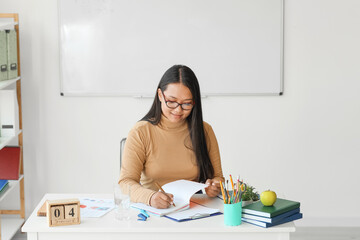 Asian female teacher sitting at table in classroom