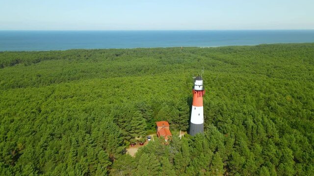 Aerial flight over Stilo lighthouse in forest trees near ocean on blue sky day