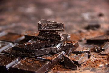 Dark chocolate bar with chocolate pieces with cocoa powder on brown background, close-up view