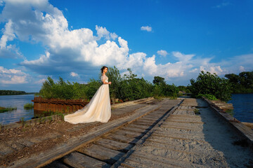beautiful girl in a white wedding dress in the summer by the old railroad against the background of a bright blue sky with clouds. The river is visible in the distance.