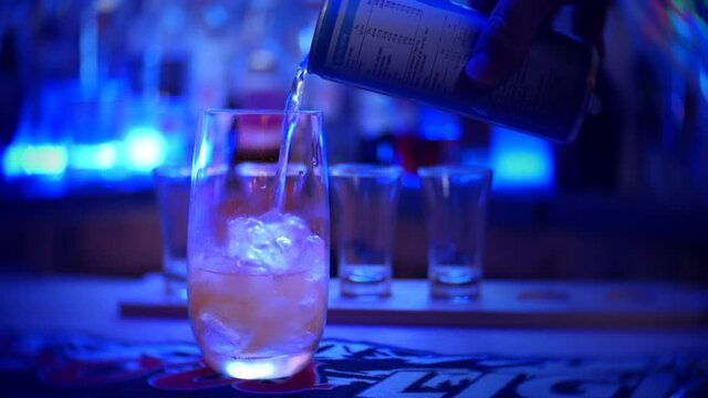 An energy drink is poured from a can into a glass filled with ice. Shot glasses can be seen on the bar in the background. Bottles of alcohol are on display at the back of the bar.