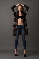 beautiful slender blonde model in a black bra, jeans on the shoulders put on a coat posing on a gray background