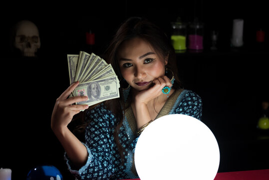 Portrait of Asian beautiful Gypsy fortune teller woman in dark room holding money bank with light illuminated crystal ball