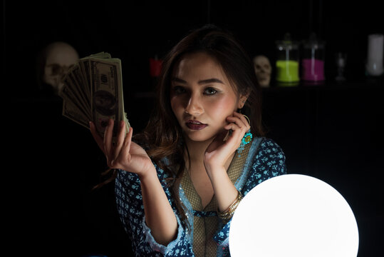 Portrait of Asian beautiful Gypsy fortune teller woman in dark room holding money bank with light illuminated crystal ball