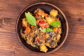 Italian vegetable stew with potatoes, eggplants, tomatoes, pepper in a rustic clay plate, decorated with fresh basil leaves on a woody background - Ciambotta, cianfotta in Neapolitan style.