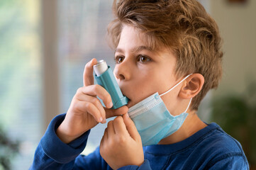 Young Boy with face mask using asthma inhaler to treat breathe issues and inflammatory disease amid Covid-19 pandemic. Difficulty Breathing in a Face Mask concept.