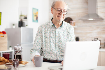 Old man in the kitchen working remotely. Daily life of senior man in kitchen during breakfast using laptop holding a cup of coffee. Elderly retired person working from home, telecommuting using remote