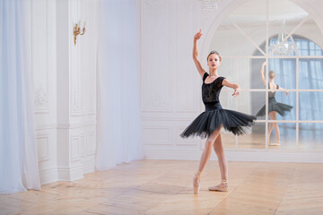 young ballerina in a black tutu is dancing on pointe in large bright hall in front of a mirror.