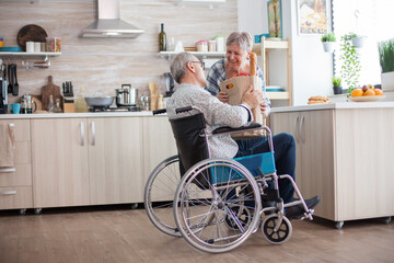 Senior woman taking grocery paper bag from handicapped husband in wheelchair. Mature people with...