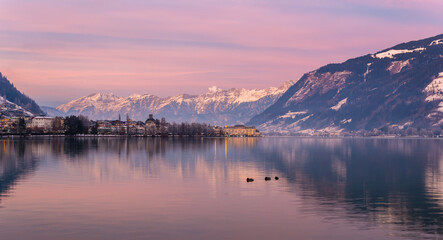 Zell am See in winter evening. View of Lake Zell, town, mountains and snow with reflections in water. Alpine town at purple dusk. Famous ski resort in Alps, Austria. Winter wonderland for travel