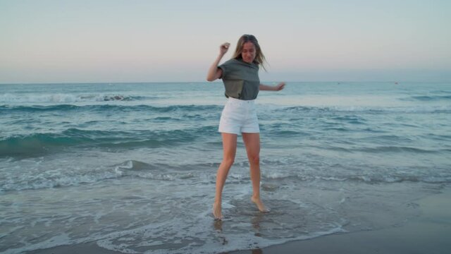 Authentic beautiful young woman dances happy in shoreline waves rolling over beach. Excited and happy, calm joyful pure happiness from being one with nature. Enjoy summertime vacation