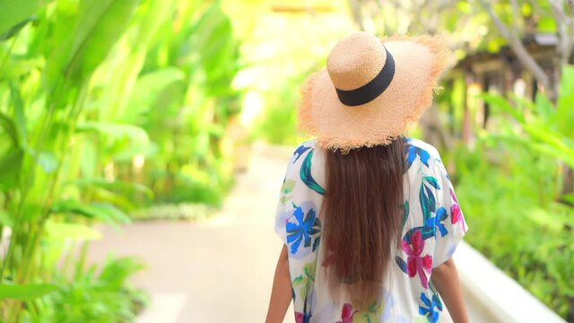 Back of stylish young female in colorful summer dress and hat walking on pathway with tropical plants on sunny day, slow motion. Holiday and fashion concept, static full frame