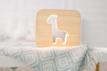 Fototapeta na wymiar Close up of stylish wooden night lamp with giraffe cut out picture, on gray blanket at cozy light bedroom interior. Wooden decorations at home interior