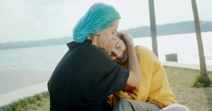 Sincere hugs playing with hair in the wind two LGBT lesbian girls sitting spending time together in nature by the lake