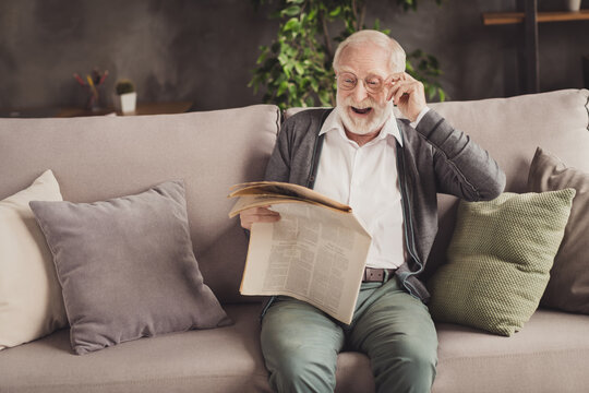 Photo of smiling pensioner wear grey cardigan sitting couch reading newspaper arm glasses indoors house flat