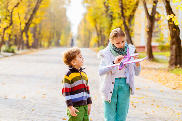 Fototapeta premium Little girl and boy,launches toy plane, into the air against background of autumn park. Children playing toy plane. Cute sister and brother, stands on track perspective, launches purple toy plane.