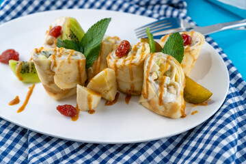 Simple homemade pancakes cut with banana, kiwi, strawberry jam on a white plate. Creamy curd filling. The food is decorated with mint. Rolled thin roll of pancake.