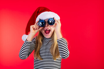 8 year old girl in santa hat covering her eyes with blue baubles. Studio photo on red background.