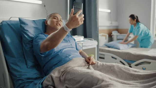 Medical Hospital Ward: Portrait of Handsome Chinese Man with Nasal Cannula Resting in Bed, Uses Smartphone to Video Chat Conference with His Family, Friends, Shooting Blog. Surgery Recovery Room.