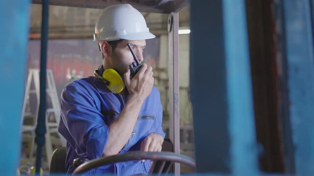 Concentrated forklift caucasian driver operating machine and holding hand on steering wheel  radio comminication operate teamwork with transportation of goods cargo in factory warehouse workshop.