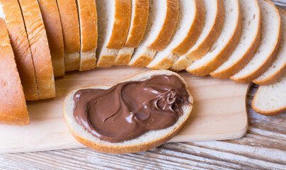 A loaf of bread and a slice of bread spread with chocolate-nut cream on a wooden background. Delicious Breakfast with chocolate and nut paste.
