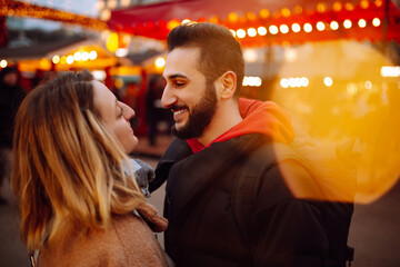 Happy couple in love walking on Christmas market at evening. Young woman and man at festive street market  enjoying winter moments. Lights around. Christmas, New Year.