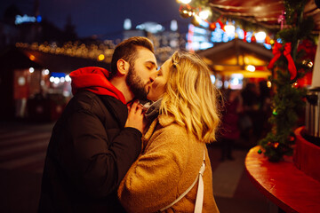 Happy couple in love walking on Christmas market at evening. Young woman and man at festive street market  enjoying winter moments. Lights around. Christmas, New Year.