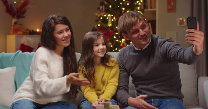 Portrait of happy Caucasian parents with little cute daughter waving hands while speaking on video call online on smartphone in decorated room with xmas tree on Christmas. New Year celebration.