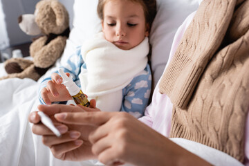 Fototapeta premium ill girl holding nasal spray near mother pointing with finger at thermometer on blurred foreground