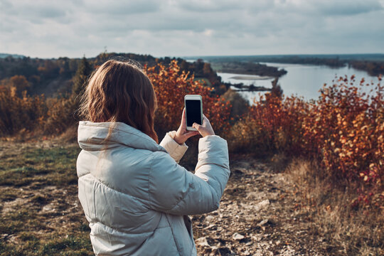 Back view of young woman taking photos of landscape with smartphone during trip on autumn sunny day