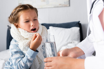 pediatrician giving glass of water to ill girl taking pill on blurred foreground