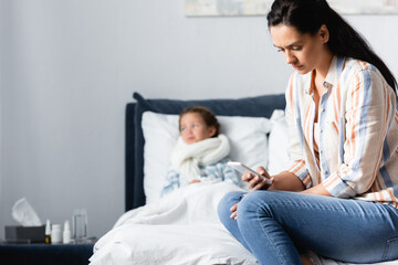 Obraz premium upset woman calling by smartphone while sitting on bed near ill daughter on blurred background