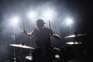 Rock band member playing drums while sitting at drum kit with backlit and smoke on background
