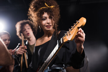 Fototapeta na wymiar Curly woman touching and looking at electric guitar, while standing near musician pointing with finger on blurred background