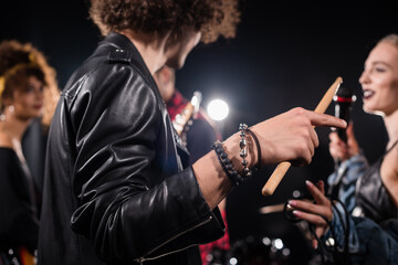 Fototapeta na wymiar Curly man with drumstick pointing with finger during rehearsal with blurred rock band musicians on background