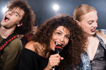 Happy rock band vocalist with microphone singing near musicians with backlit on blurred background