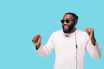 Cheerful young African American handsome man in eyeglasses enjoy favourite playlist in headphones, listens audio in music app and dance against blue isolated background, he have fun