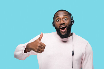 Portrait of cheerful african american call center worker in headphones with microphone standing on isolated blue background, surprised looks at camera and showing thumb up
