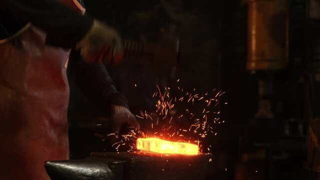 Medium shot of a man with a hammer hitting red-hot metal. A blacksmith works with metal in a forge. Sparks from impacts to metal