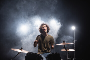 Excited curly drummer with open mouth sitting at drum kit with smoke and backlit on black