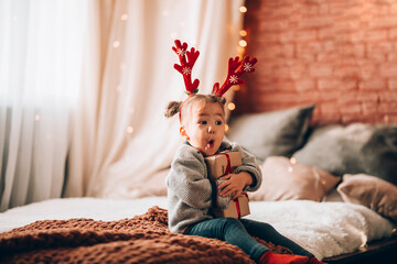 A little girl holding a large gift in a paper box on a large bed. The child has antlers on his...