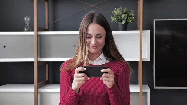 Gambling young woman girl 20s years old in burgundy sweater posing using play mobile cell phone hold gadget smartphone video pc games on modern internrt background. People lifestyle concept