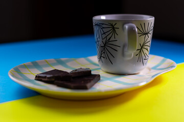 Side view of a white cup on a plate with blue and yellow lines with some chocolate portions. All of this on a blue and yellow surface with a black background.