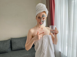 young woman with a towel on her head and a gray clay mask on her face points to a glass of clear water.