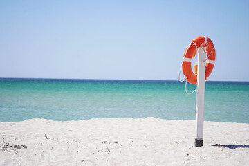 an orange lifebuoy hangs from a pole on a sandy beach against a blue sky and azure sea.