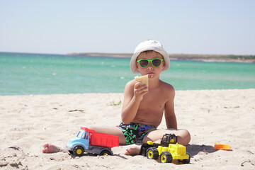 on a Sunny hot summer day, a boy plays with sand and toys, cars on the beach. a child eats ice cream while sitting on a sandy beach with an azure sea background.
