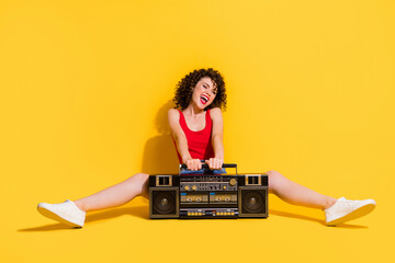 Portrait photo of pretty laughing curly girl sitting listening to music smiling happily entertaining keeping hands touching boombox isolated on vivid yellow color background